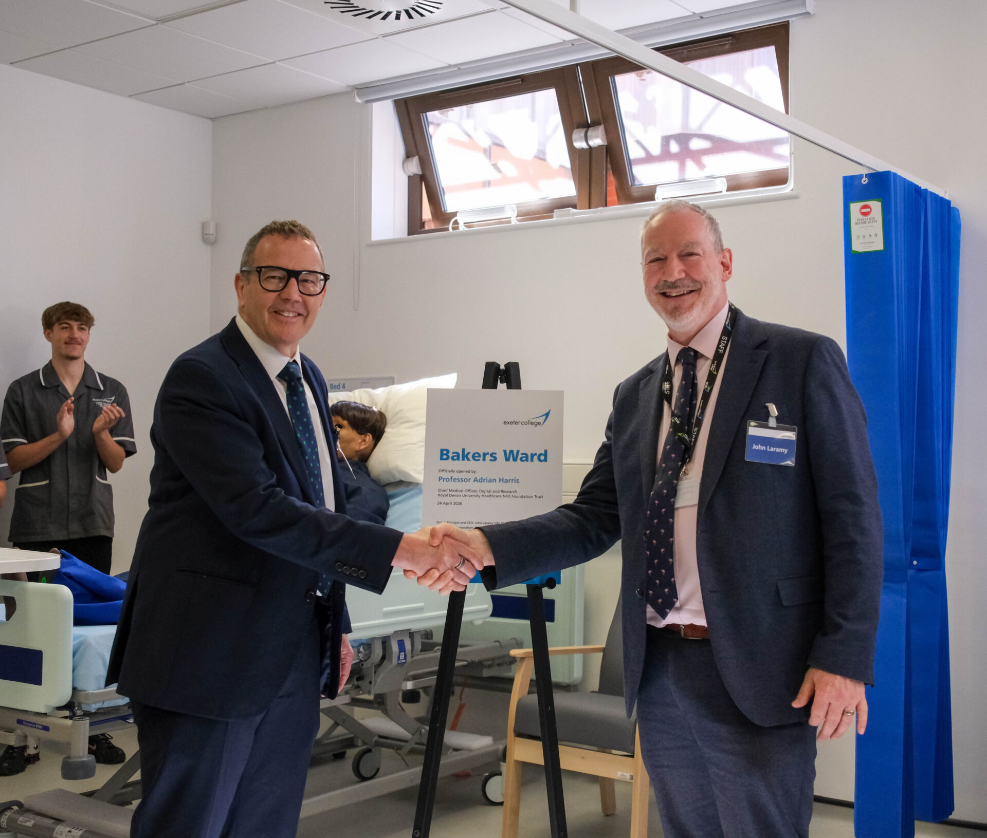 Two people shake hands in front of a plaque at Exeter College's new hospital training ward.